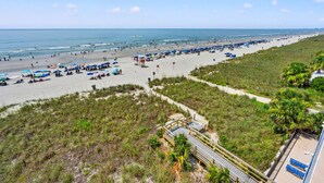 Una playa con numerosos paraguas, gente, un paseo marítimo y una zona verde con árboles.