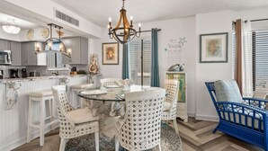 Un área de comedor con una mesa redonda de vidrio rodeada por sillas de mimbre blanco, una araña y una silla azul en la esquina.