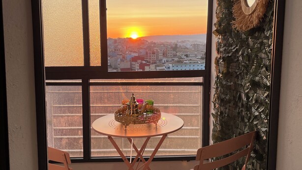 A balcony with a view of a sunset over a cityscape, a round table with a decorative item, and two wooden chairs. - Flat in tanger : wifi, prvt parking, air-co netflix... (Tanger)