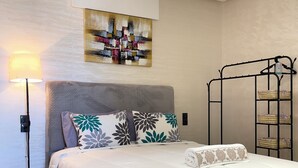 A neatly made bed with a white duvet and floral pillows, a floor lamp, a painting on the wall, and a metal shelf unit with baskets. - Flat in tanger : wifi, prvt parking, air-co netflix... (Tanger)