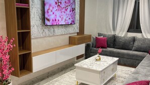 A modern living room with a large flat-screen TV mounted on a marble wall, a grey sofa with red cushions, a white coffee table, and a wooden shelving unit. - Flat in tanger : wifi, prvt parking, air-co netflix... (Tanger)