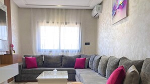 A modern living room with a grey sectional sofa, a white coffee table, and a round ottoman. There is a large window with sheer curtains, a ceiling light fixture, and a wall-mounted air conditioner. - Flat in tanger : wifi, prvt parking, air-co netflix... (Tanger)