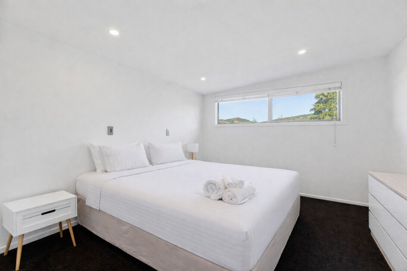 A modern, minimalist bedroom with a large bed, a bedside table, a nightstand, and a window with a view of greenery.