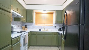 Une cuisine de taille réduite avec des meubles verts, un réfrigérateur noir, une cuisinière blanche et une fenêtre équipée de stores.