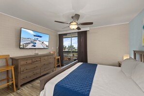 A hotel room with a bed, a TV mounted on the wall, a ceiling fan, a wooden dresser, and a view of the outdoors. - Beachfront Family-Friendly Condo w/ Heated Pool! (South Padre Island)