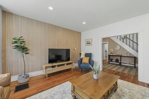 A modern living room with a flat-screen TV, a blue armchair, a wooden coffee table, and a staircase leading to an upper level. - Spacious Central Berkshires Home for Groups (Lee)