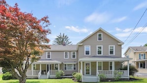 A two-story residential house with a front porch, a small garden, and a tree with autumn leaves. - Spacious Central Berkshires Home for Groups (Lee)