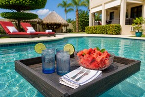 Ein Tablett mit Getränken und Wassermelone am Pool, Liegestühle und im Hintergrund ein Gebäude mit einem Reetdach.