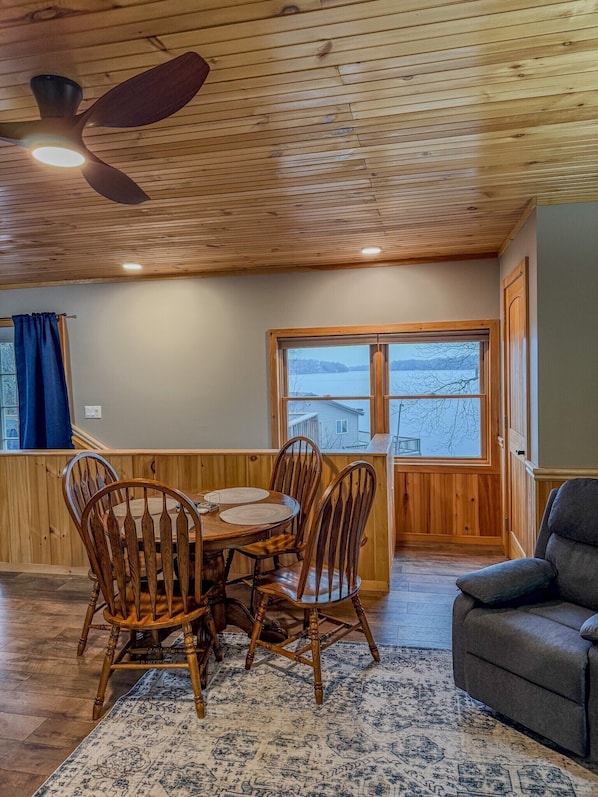 A wooden-paneled room with a dining table, chairs, a ceiling fan, and a large window with a view of a lake. - Lakeside Loft on Black Lake (Hammond)