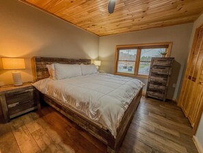 A bedroom with a wooden ceiling, a bed with white bedding, a nightstand with a lamp, a dresser, and a window with a view of the outside.