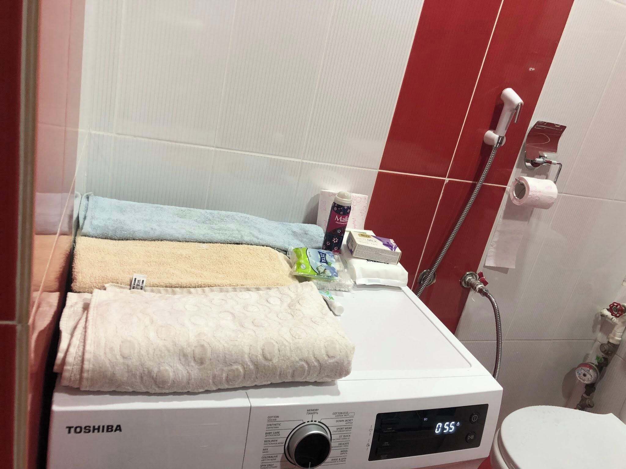 Une machine à laver Toshiba dans une salle de bain avec un mur carrelé en rouge et blanc, des toilettes et un espace douche.