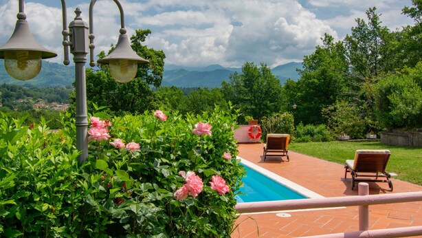 Zona de piscina con vistas a montañas, rodeada de vegetación exuberante y algunas sillas.