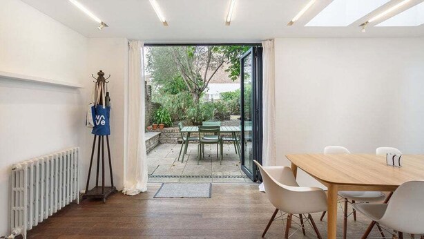 A modern room with a wooden floor, a dining table with chairs, a radiator, and a view of an outdoor garden through a glass door.