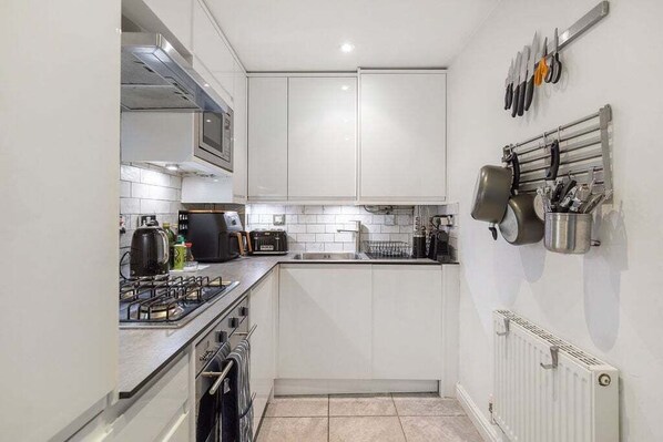 Een kleine keuken met witte kastjes, een gasfornuis, een magnetron en een waterkoker. Bestek hangt aan de muur en er is een afwassigrek.
