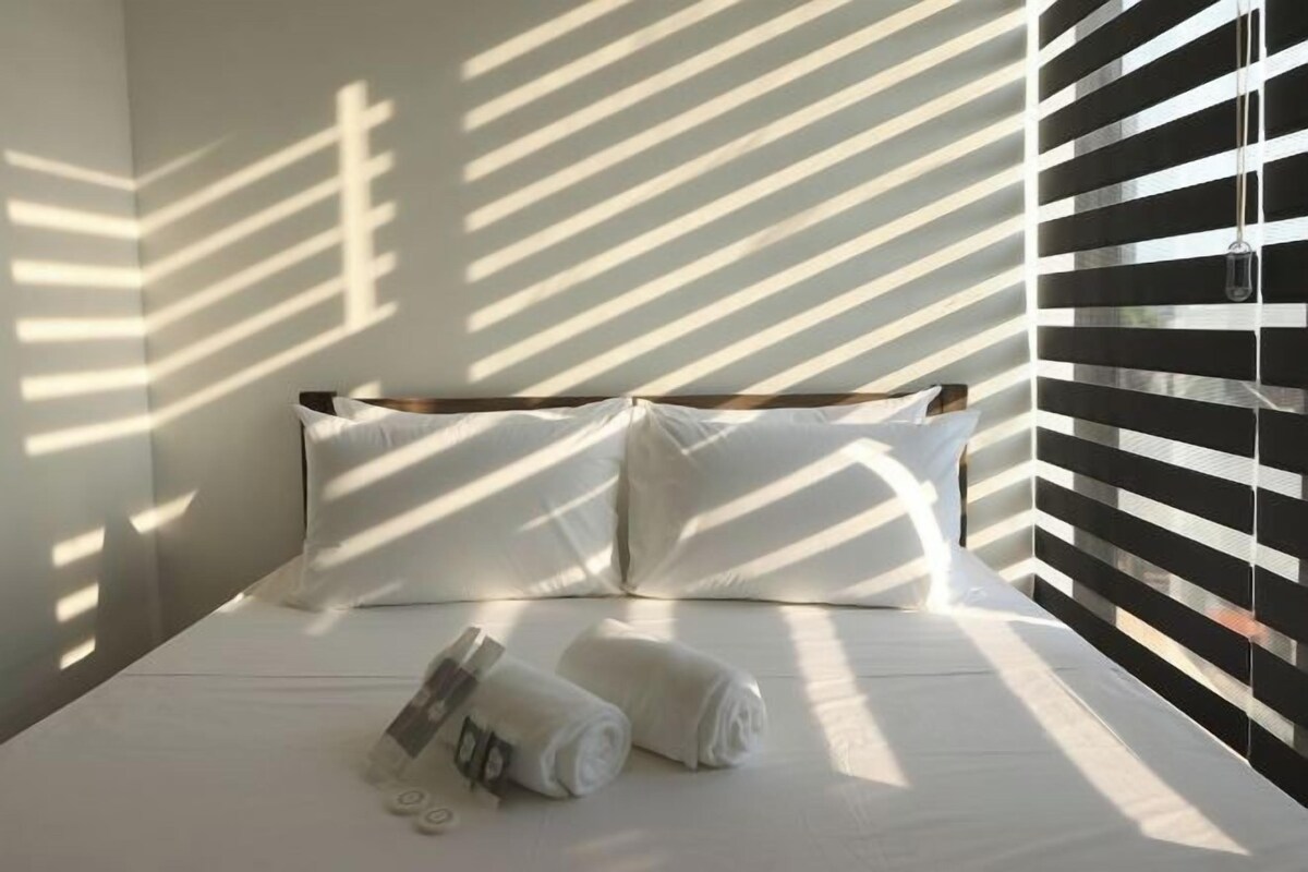 A neatly made bed with white linens and pillows, a towel, and a small bottle, with sunlight casting shadows on the wall.