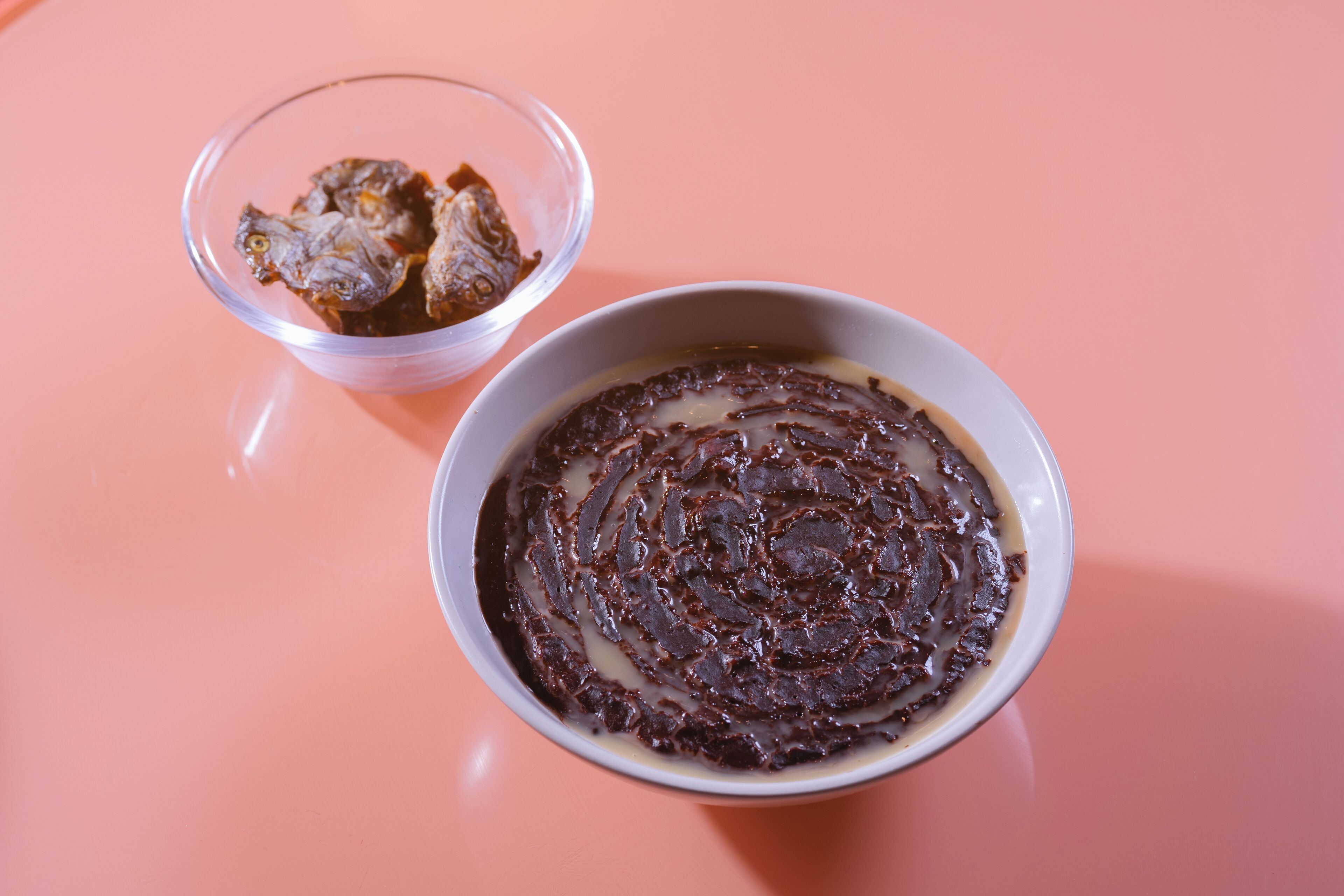 Un bol de postre de chocolate junto a un vaso de una bebida oscura sobre una superficie rosa.