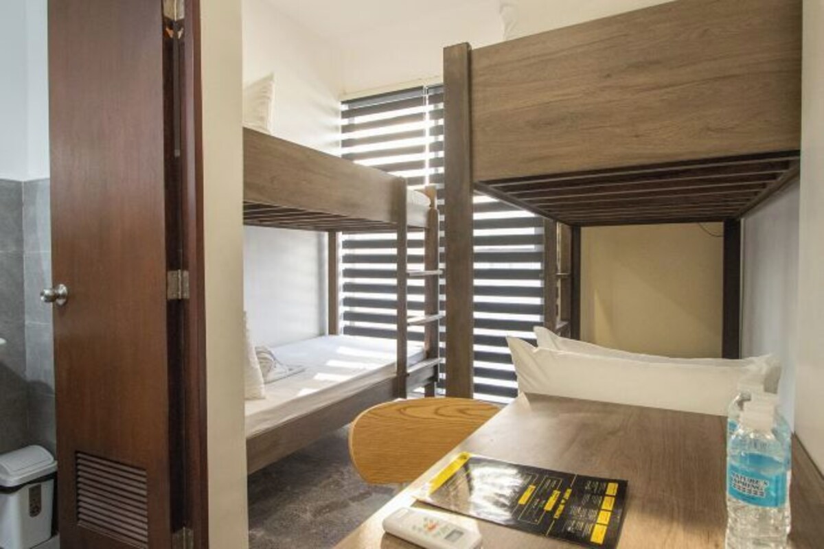 A bunk bed room with a wooden door, a small table with a book and a bottle, and a window with blinds.