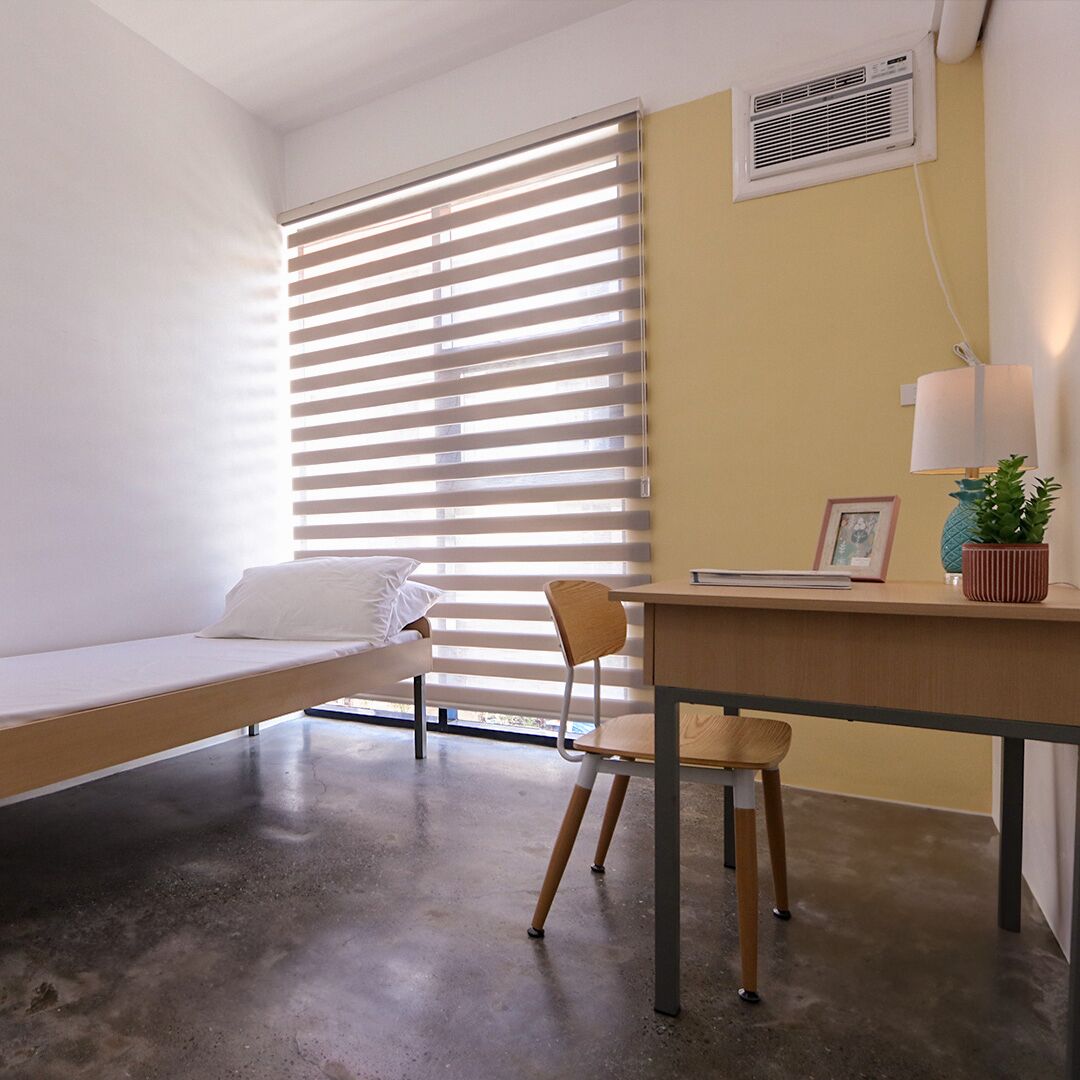 A room with a bed, a desk, a chair, a window with blinds, a wall-mounted air conditioner, and a lamp.