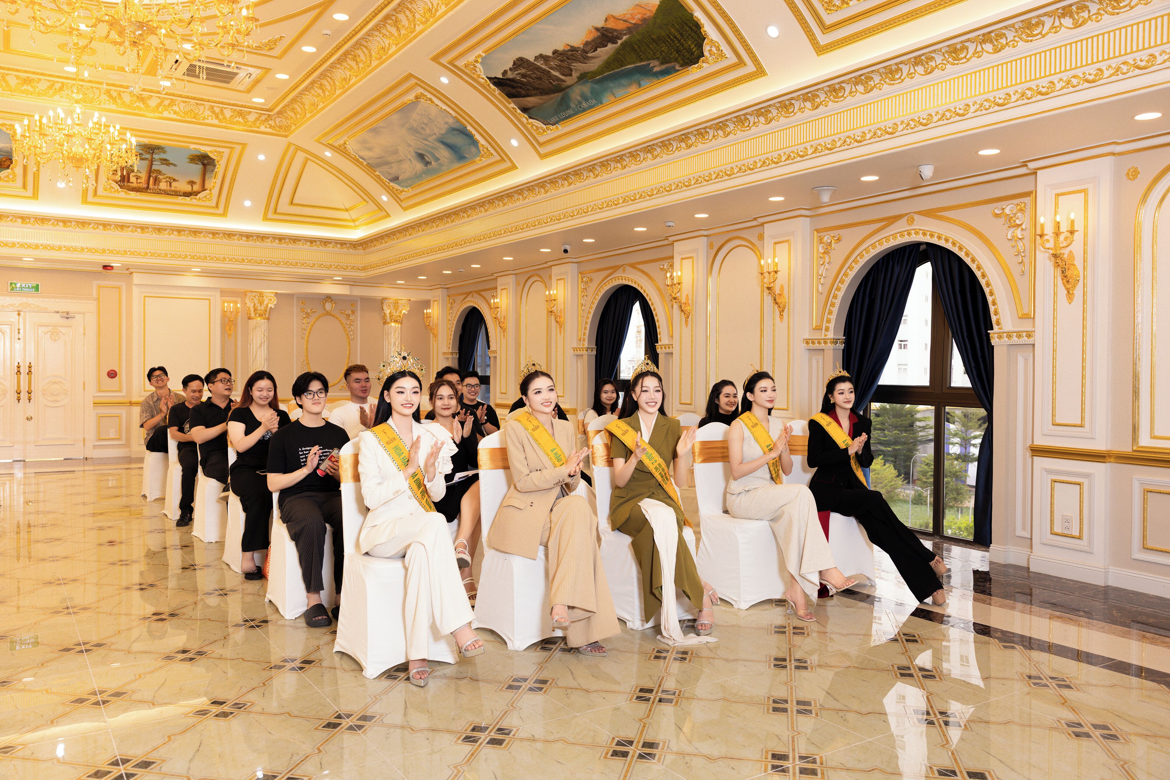 A group of people seated in a luxurious room with ornate gold detailing, large windows, and a chandelier.