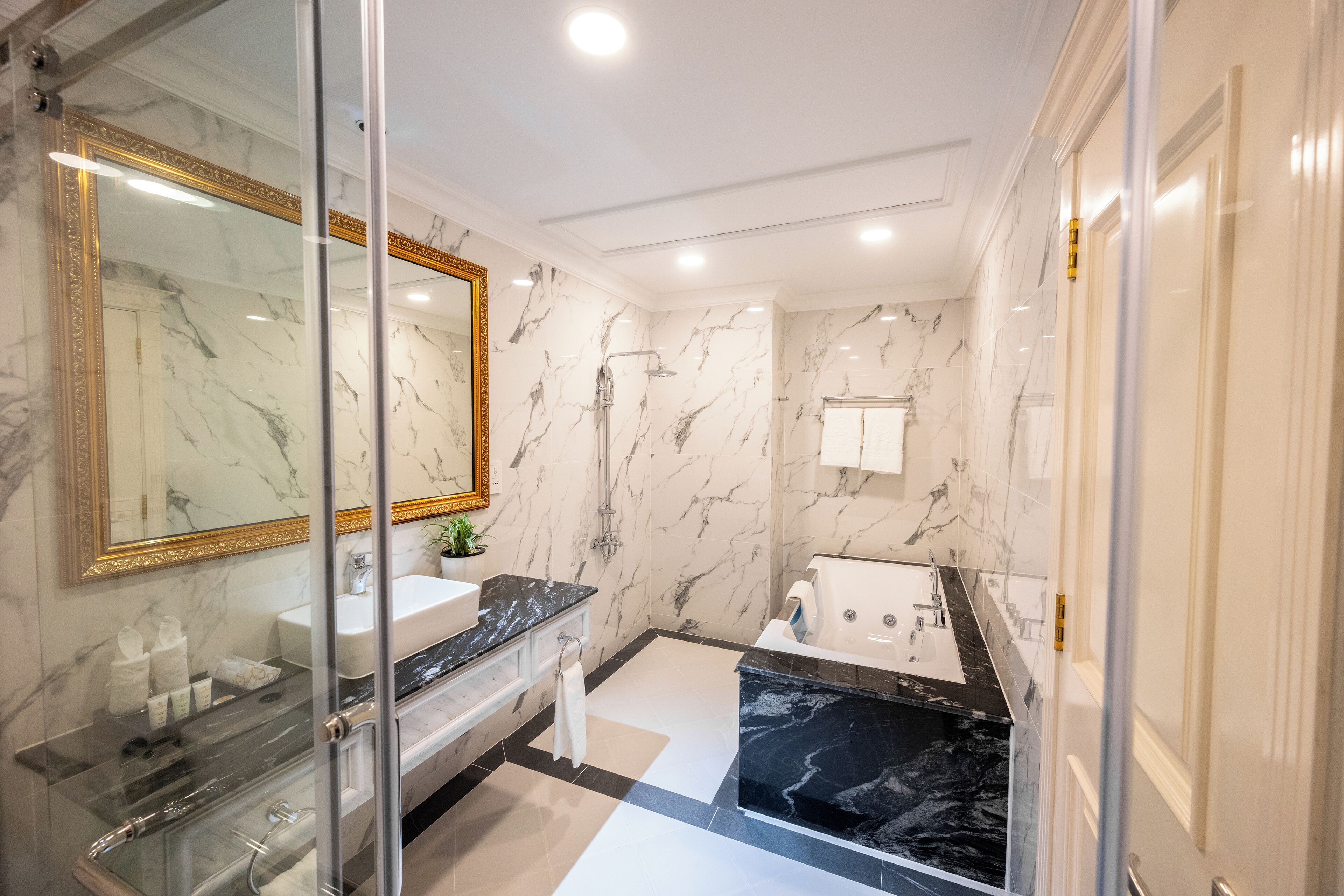 Un baño lujoso con paredes de mármol, un espejo grande, cabina de ducha con vidrio y una bañera de mármol negro, además de un lavamanos blanco.