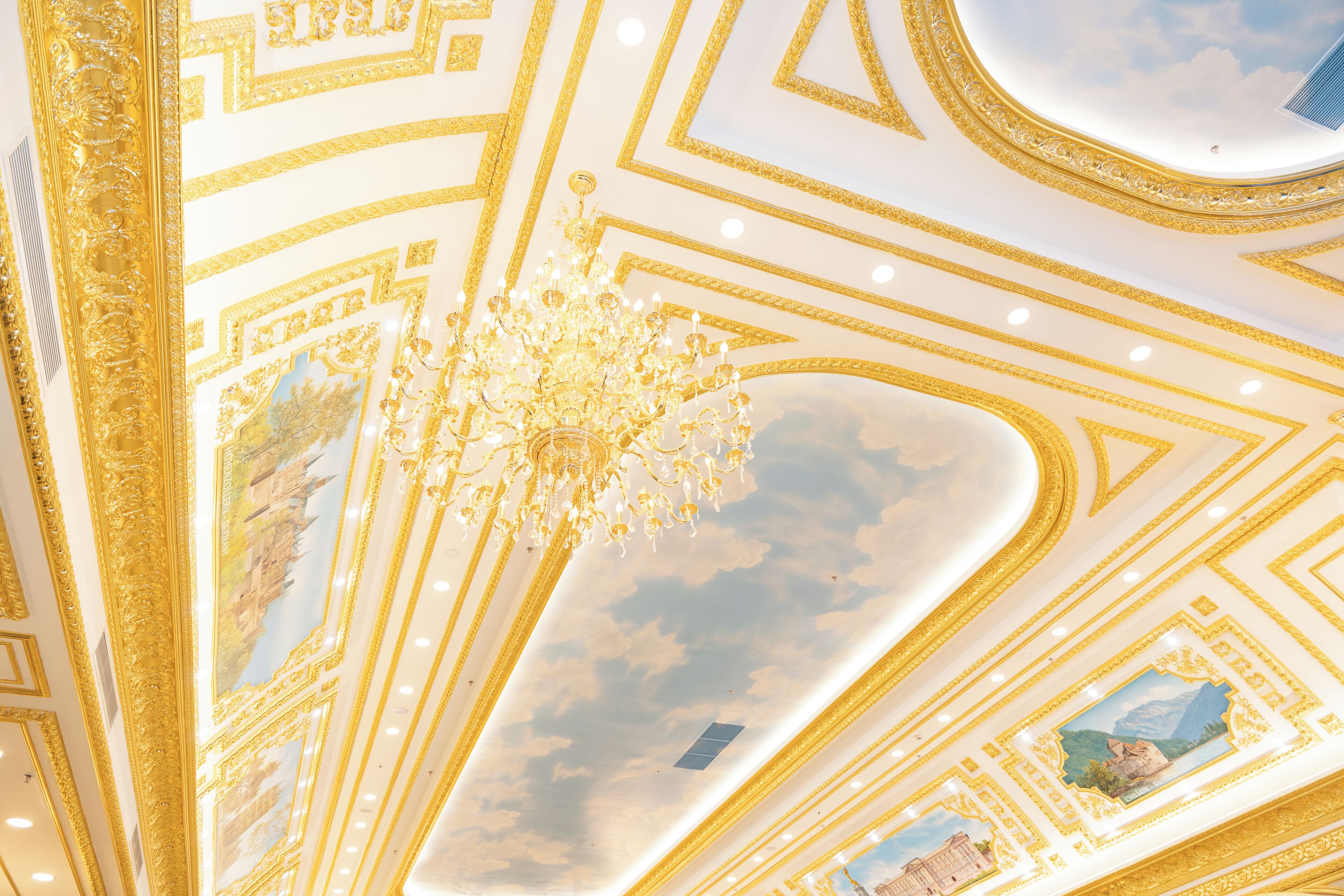 A ceiling with gold and white geometric patterns, a chandelier, and a skylight with a painted sky.