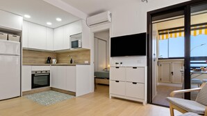 A compact kitchen with white cabinets, a microwave, and a built-in oven. There is a flat-screen TV mounted above a cabinet, and a sliding glass door leading to a balcony with outdoor seating. - Montemar Puerto - Beachfront, Terrace, Swimming Pool. Sea Views (Torremolinos)