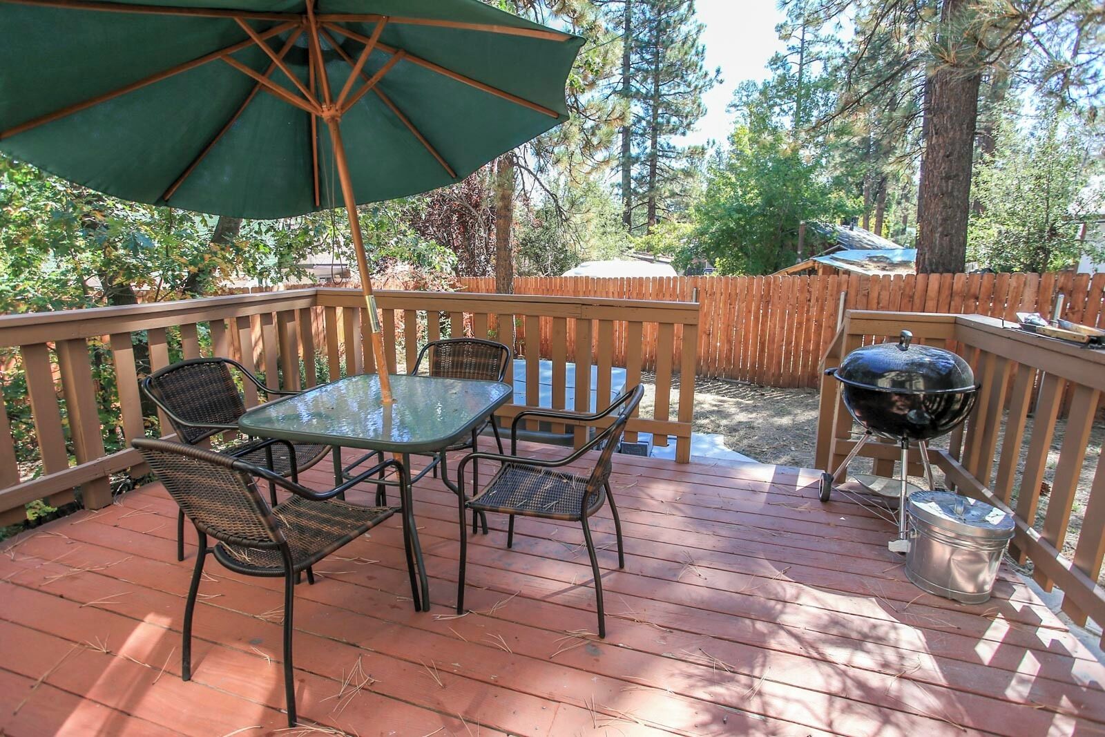 A wooden deck with a glass table and chairs, a barbecue grill, and a green umbrella.