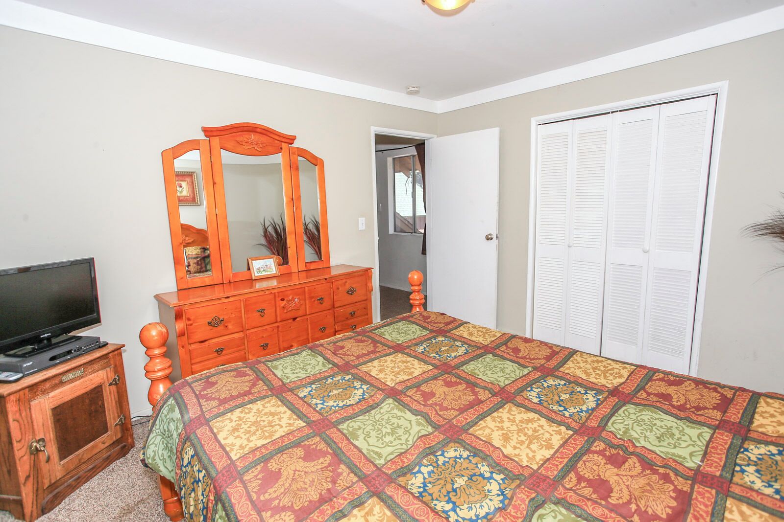 A bedroom with a large bed, a wooden dresser, a television on a cabinet, a mirror, and a closet with white shutters.