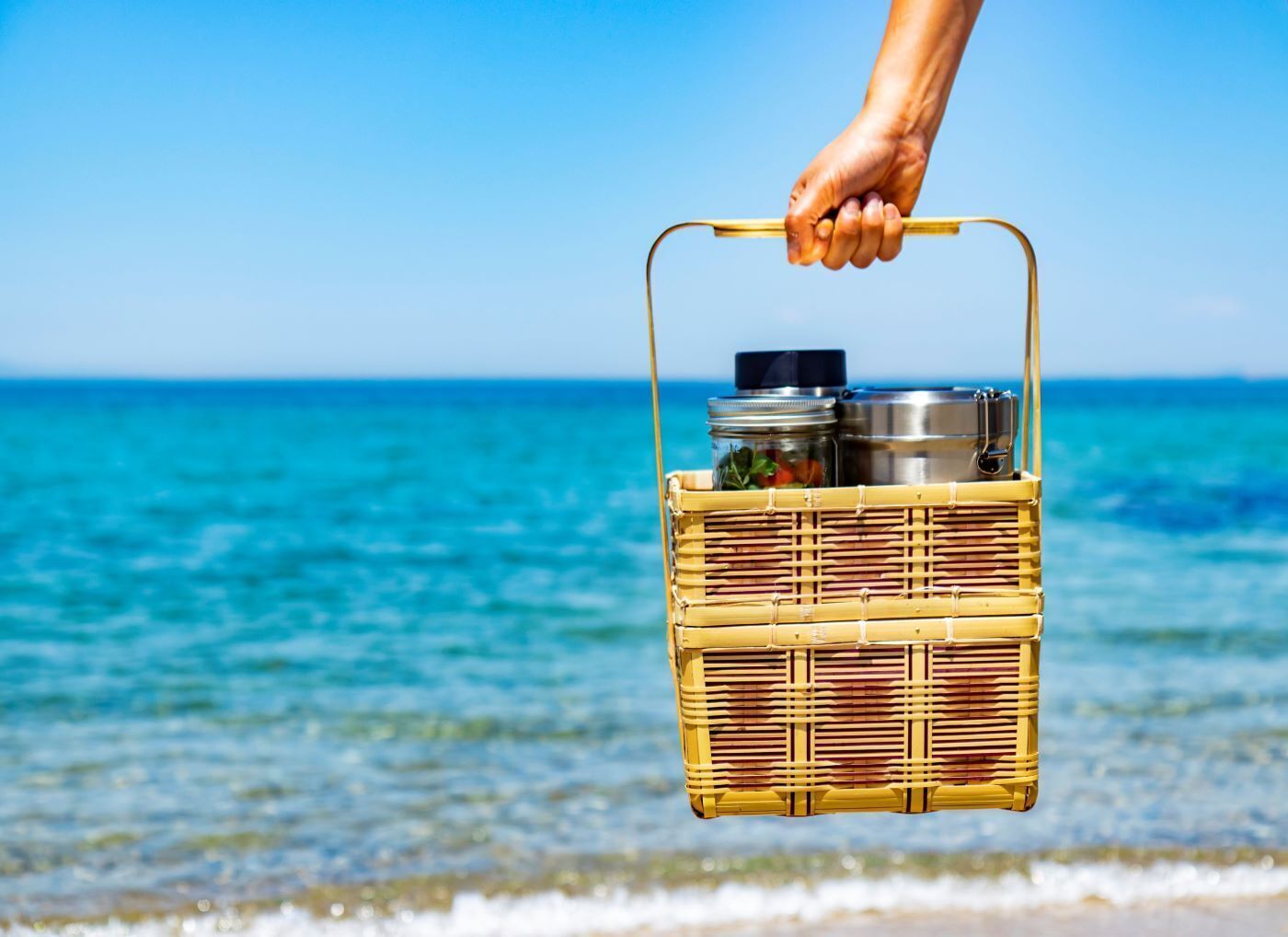 Una mano sosteniendo una cesta de mimbre con un recipiente metálico y un tarro, sobre una playa con agua azul cristalina.