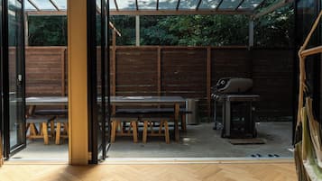 A modern, open-plan space with a wooden floor, a glass enclosure, a wooden table with benches, and a barbecue grill.