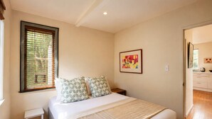 A bedroom with a bed, a small bedside table, a window with blinds, a framed picture on the wall, and a view into the adjacent room. - Beachlovers Onetangi - Walk to Onetangi Beach (Waiheke Island)
