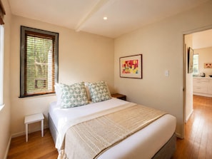 Un dormitorio con cama, una mesita de noche pequeña, ventana con persianas, un cuadro en la pared y vista a la habitación contigua.