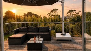 A balcony with a sectional sofa, a small table with a bottle and glasses, and a large umbrella. - Beachlovers Onetangi - Walk to Onetangi Beach (Waiheke Island)