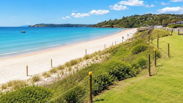 A beach with a clear view of the ocean, a boat in the water, and a grassy area with a fence. - Beachlovers Onetangi - Walk to Onetangi Beach (Waiheke Island)