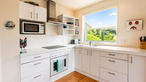 A modern kitchen with white cabinets, a microwave, an oven, and a window with a view of greenery. - Beachlovers Onetangi - Walk to Onetangi Beach (Waiheke Island)
