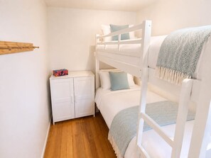 Ein Stockbett mit weißem Kopfteil und einem unteren Bett mit Kissen. Es befindet sich ein kleiner weißer Schrank mit einem farbigen Gegenstand darauf.