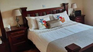 Un lit bien fait, agrémenté de coussins à motifs floraux, d’une tête de lit en bois et d’une table de chevet avec une lampe.