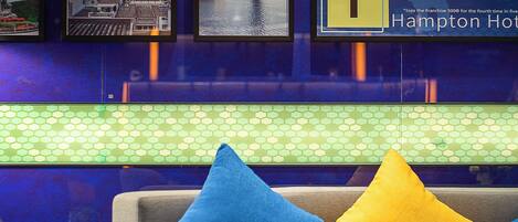 A hotel lobby with framed pictures on the wall, a seating area with blue and yellow pillows, and a green patterned tile backsplash.