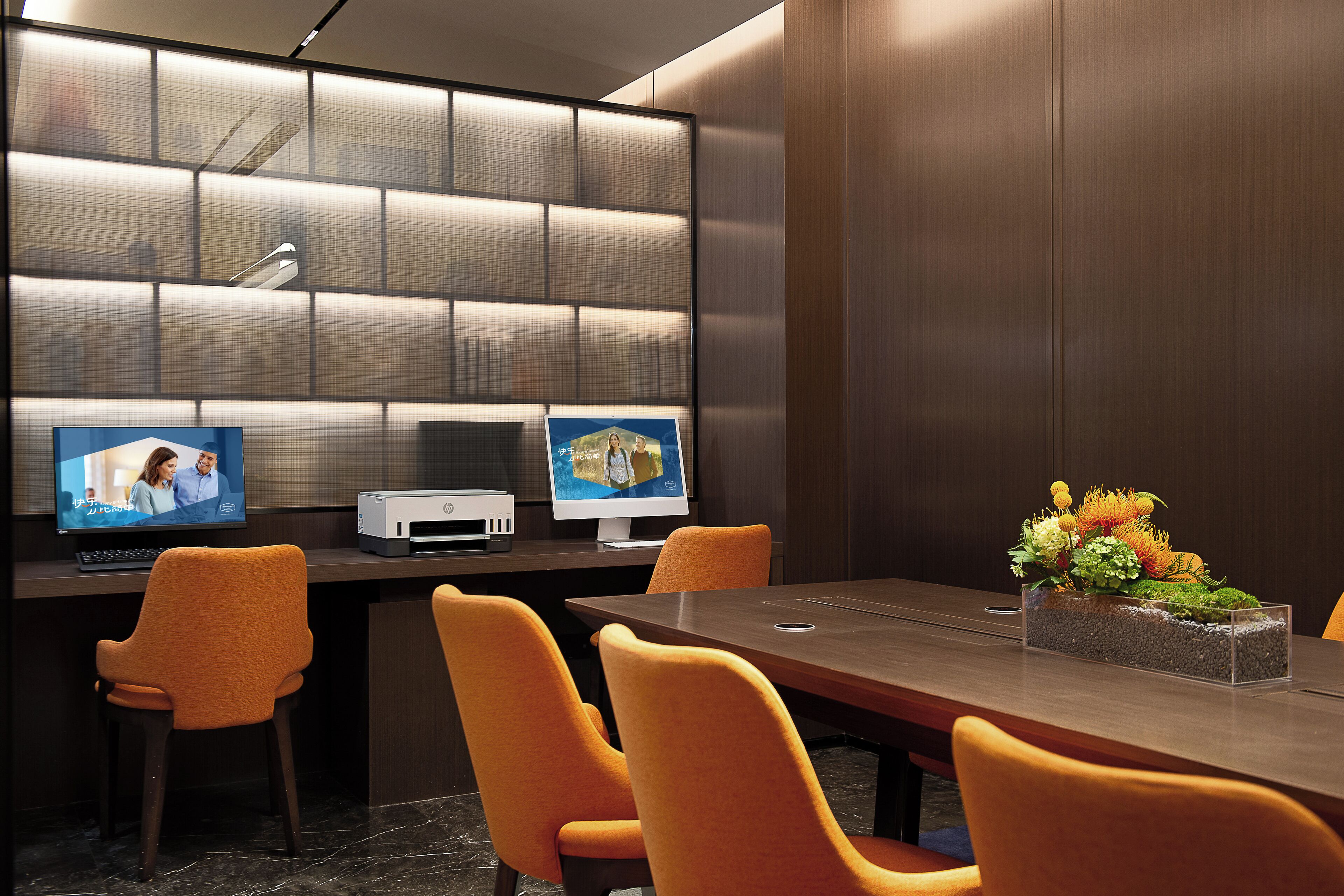 A conference room with a long table, four orange chairs, two monitors, and a floral arrangement.