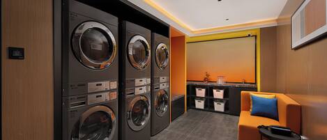 A modern laundry room with three stacked washers and dryers, a small seating area with a sofa and armchair, and a kitchen counter in the background.