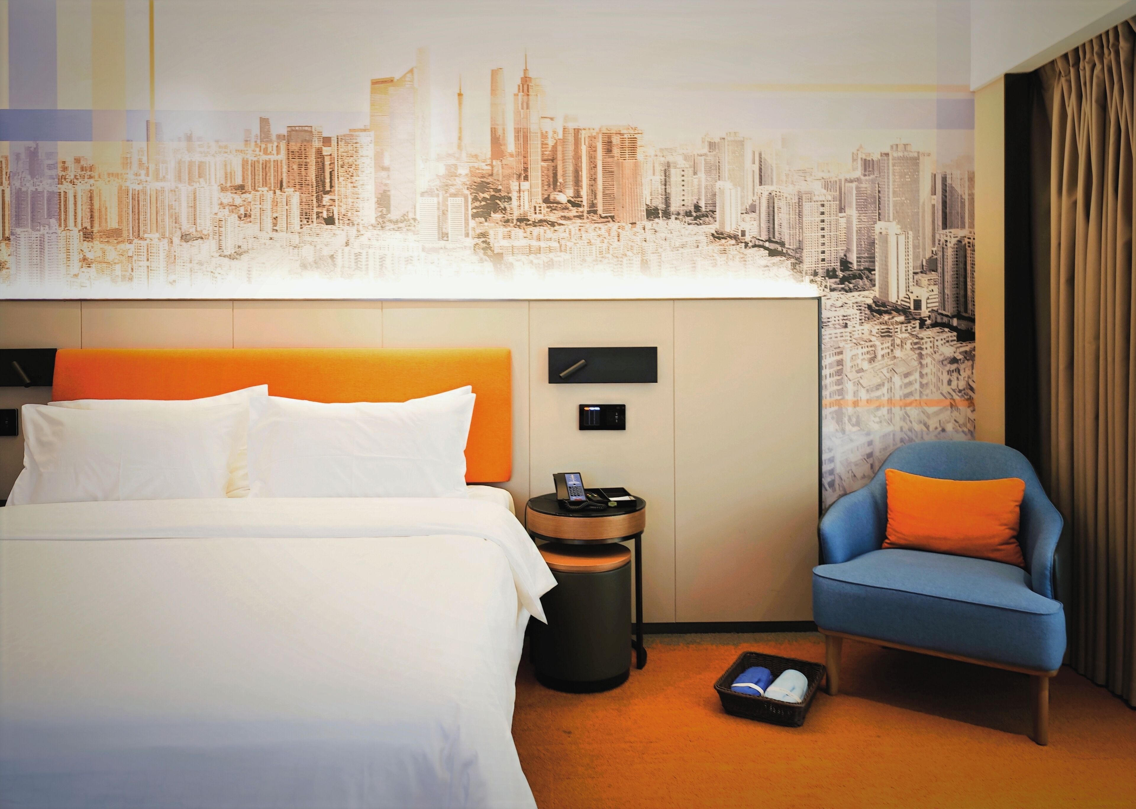 Habitación de hotel con cama, una silla, una mesita de noche y un mural de la ciudad en la pared.