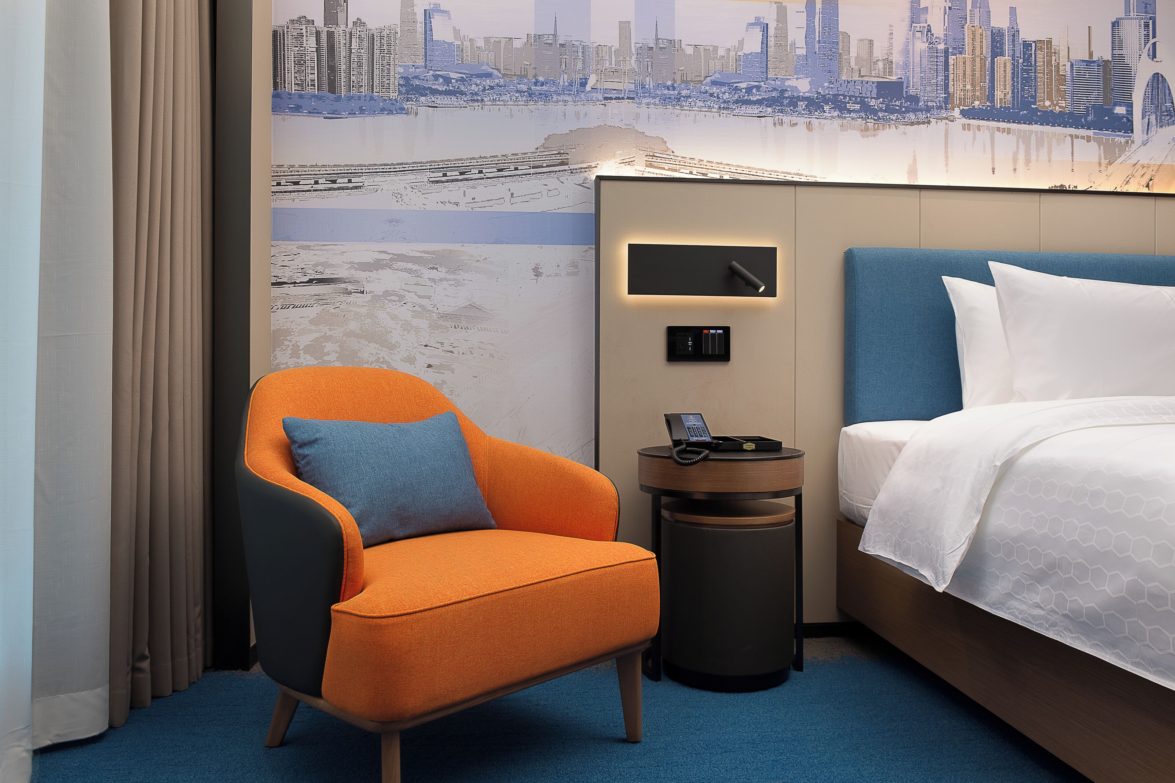 Habitación de hotel con un mural de la ciudad, un cabecero azul, un sillón naranja, una mesita de noche con un teléfono y una cama bien hecha.