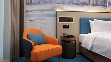 A hotel room with a cityscape mural, a blue headboard, an orange armchair, a bedside table with a phone, and a neatly made bed.