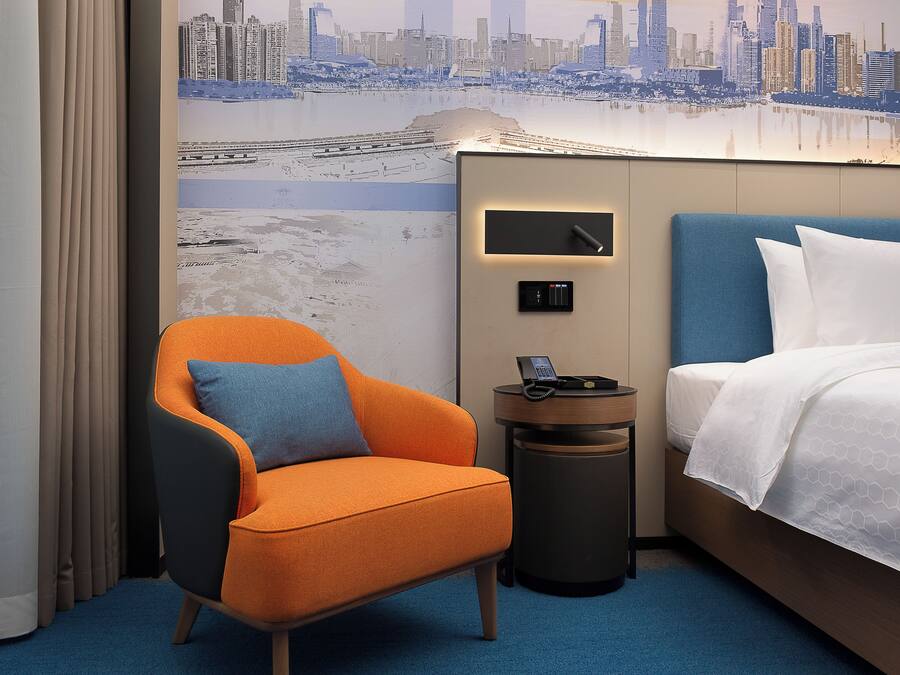 A hotel room with a cityscape mural, a blue headboard, an orange armchair, a bedside table with a phone, and a neatly made bed.