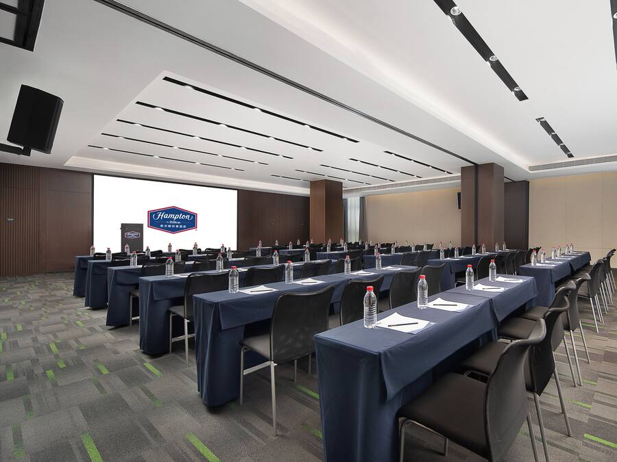 Un salón de conferencias con varias mesas, sillas y una pantalla grande que muestra el logotipo de Hampton Inn.
