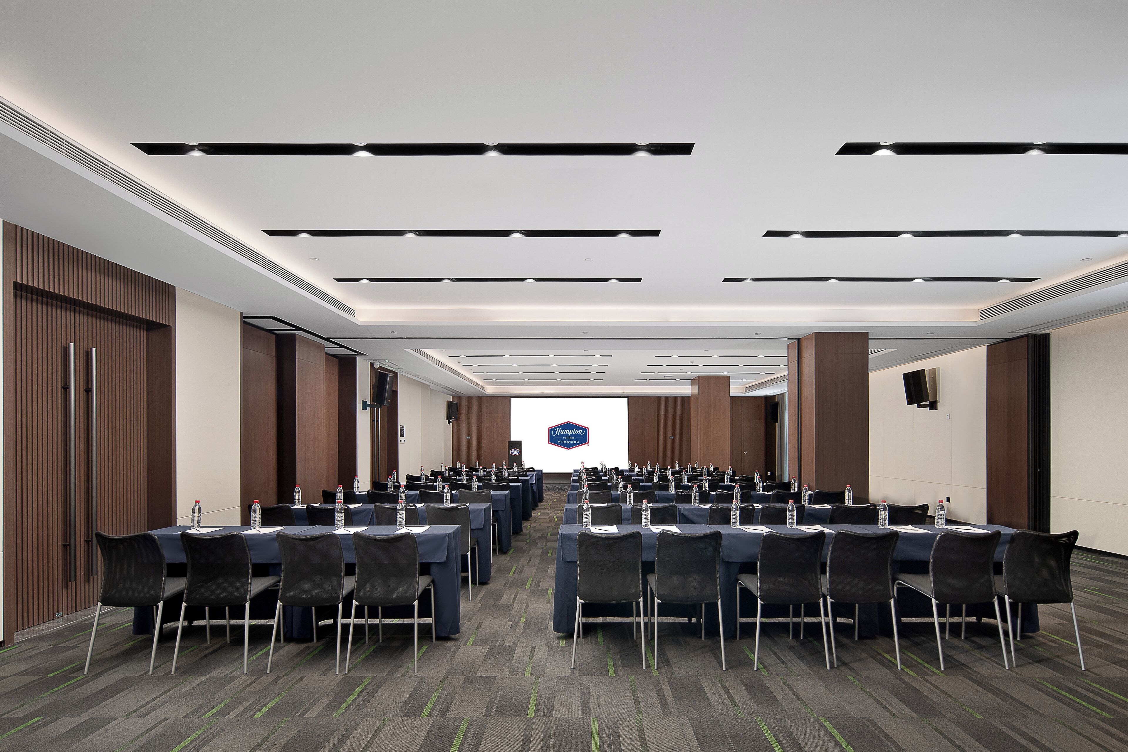 A conference room with rows of tables and chairs, a large screen at the front, and a logo on the wall.