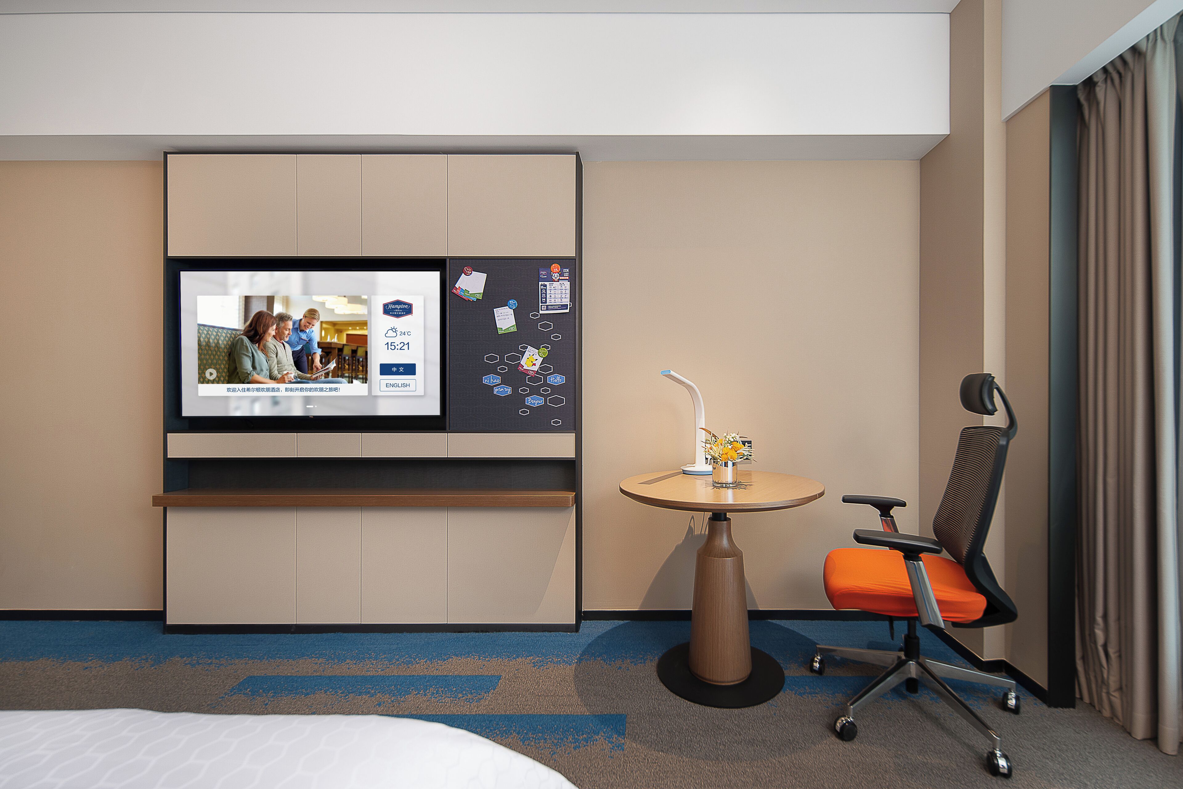 Une chambre d’hôtel comprenant un lit, un téléviseur fixé au mur, un bureau avec une chaise et une petite table sur laquelle est posé un vase.
