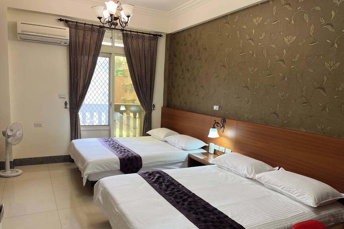 A hotel room with two beds, a ceiling fan, an air conditioner, a nightstand with a lamp, and a view of the outside balcony.