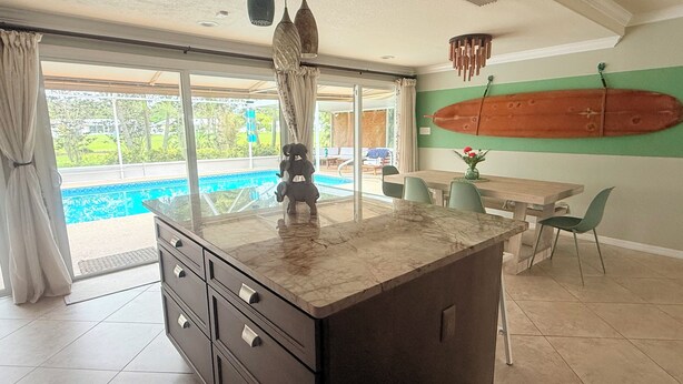 A kitchen with a marble countertop, a wooden dining table, and chairs. There is a large window with curtains, a pool visible outside, and a surfboard on the wall.