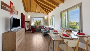 A modern living room with a TV, sofa, dining table, and large windows offering a view of the outdoors. - Viola Apartment Lake Maggiore Retreat w/ Private Garden & Parking, Casalzuigno, Italy (Casalzuigno)